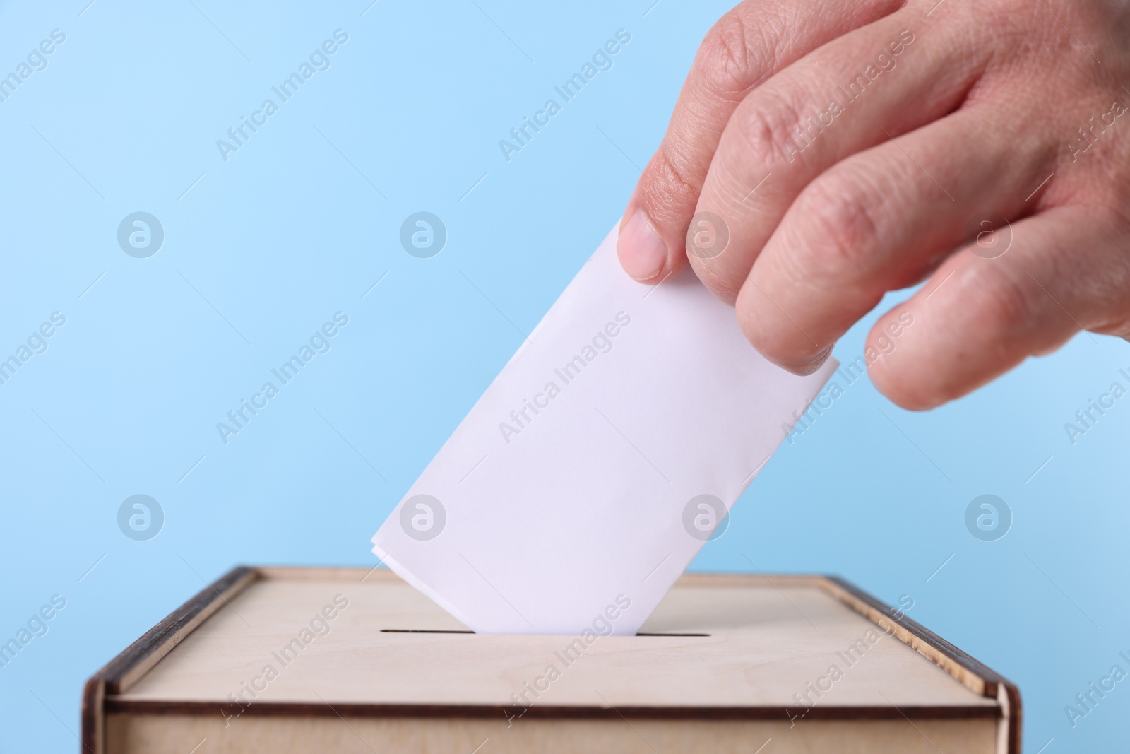 Photo of Man putting his vote into ballot box against light blue background, closeup