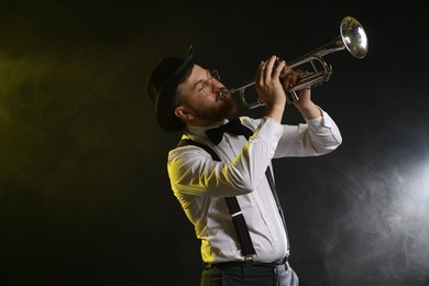 Photo of Professional musician playing trumpet on black background in color lights and smoke