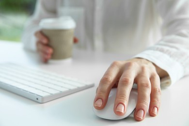 Woman with wireless mouse and paper cup at white table indoors, closeup