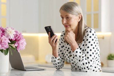 Senior woman using mobile phone at white table indoors
