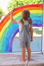 Photo of Little girl drawing rainbow on window indoors, back view