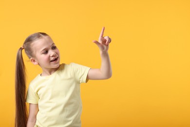Cute little girl pointing at something on orange background, space for text