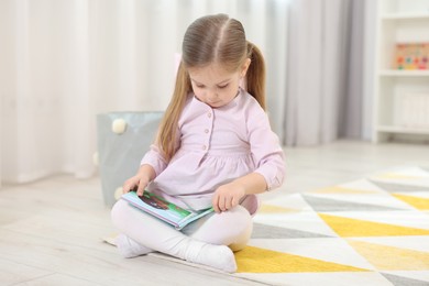 Photo of Cute little girl reading book on floor in kindergarten