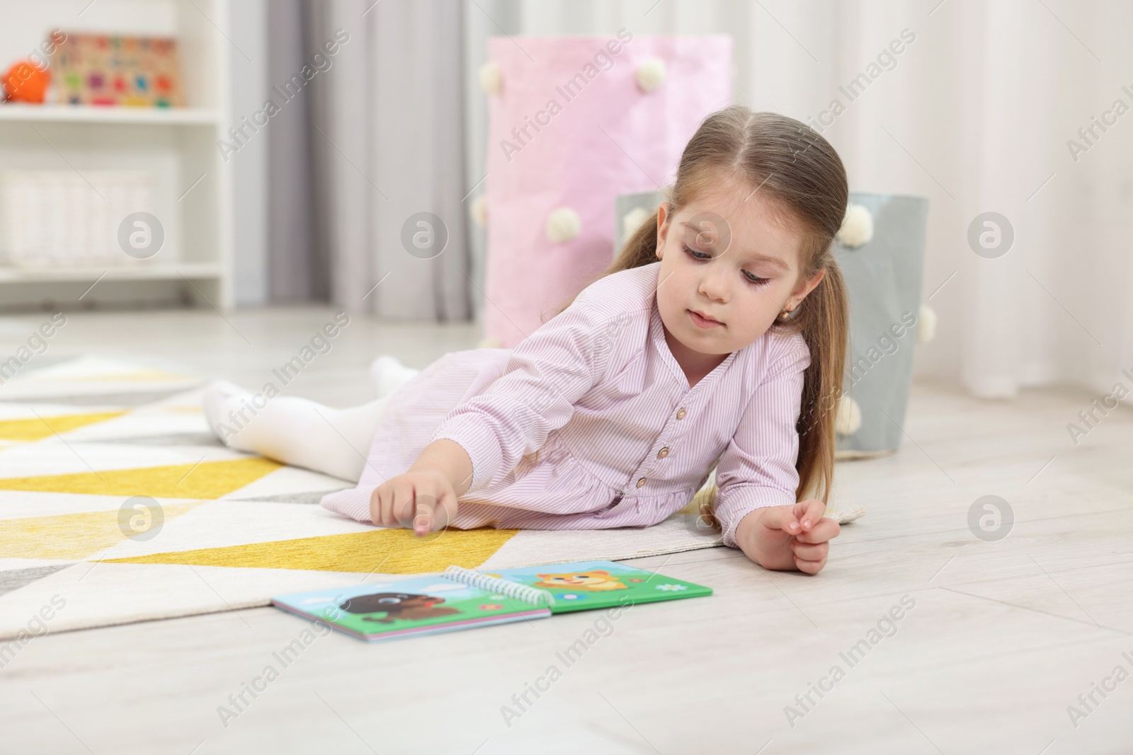 Photo of Cute little girl reading book on floor in kindergarten