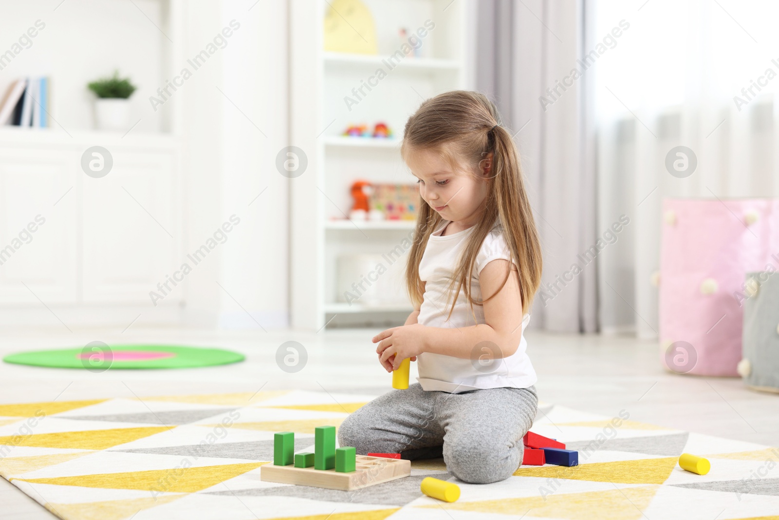 Photo of Cute little girl playing with set of wooden geometric figures on floor in kindergarten. Space for text