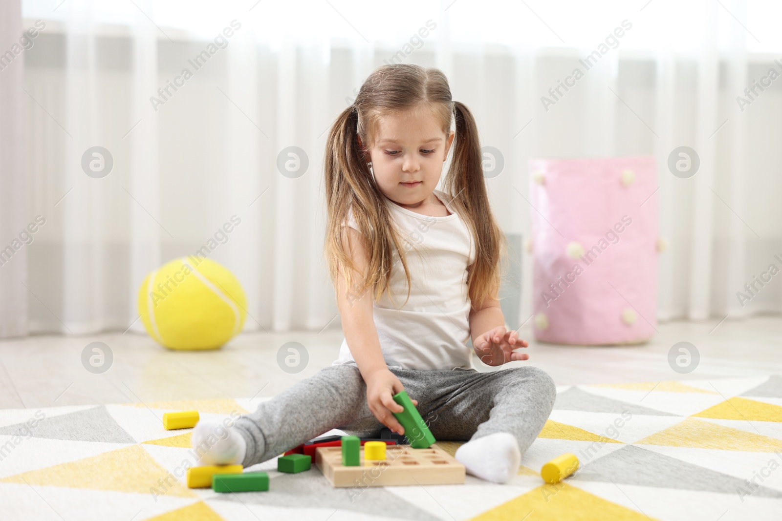 Photo of Cute little girl playing with set of wooden geometric figures on floor in kindergarten