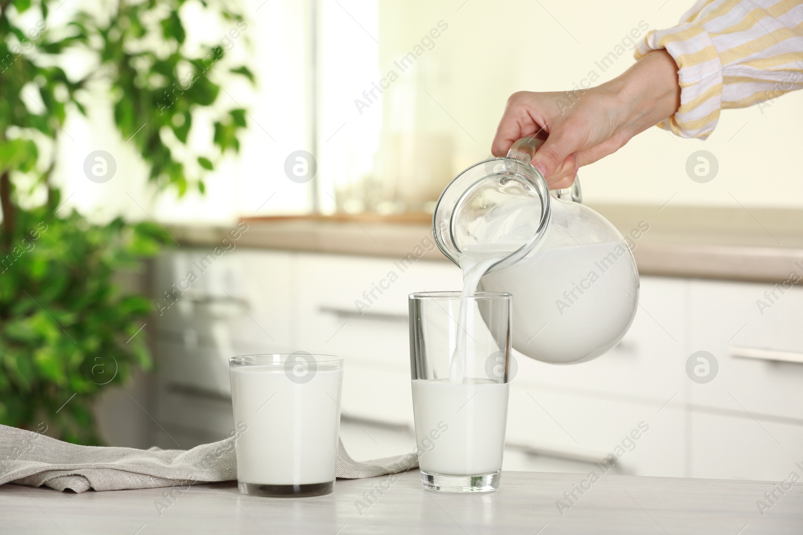 Photo of Woman pouring fresh milk from jug into glass at light wooden table in kitchen, closeup