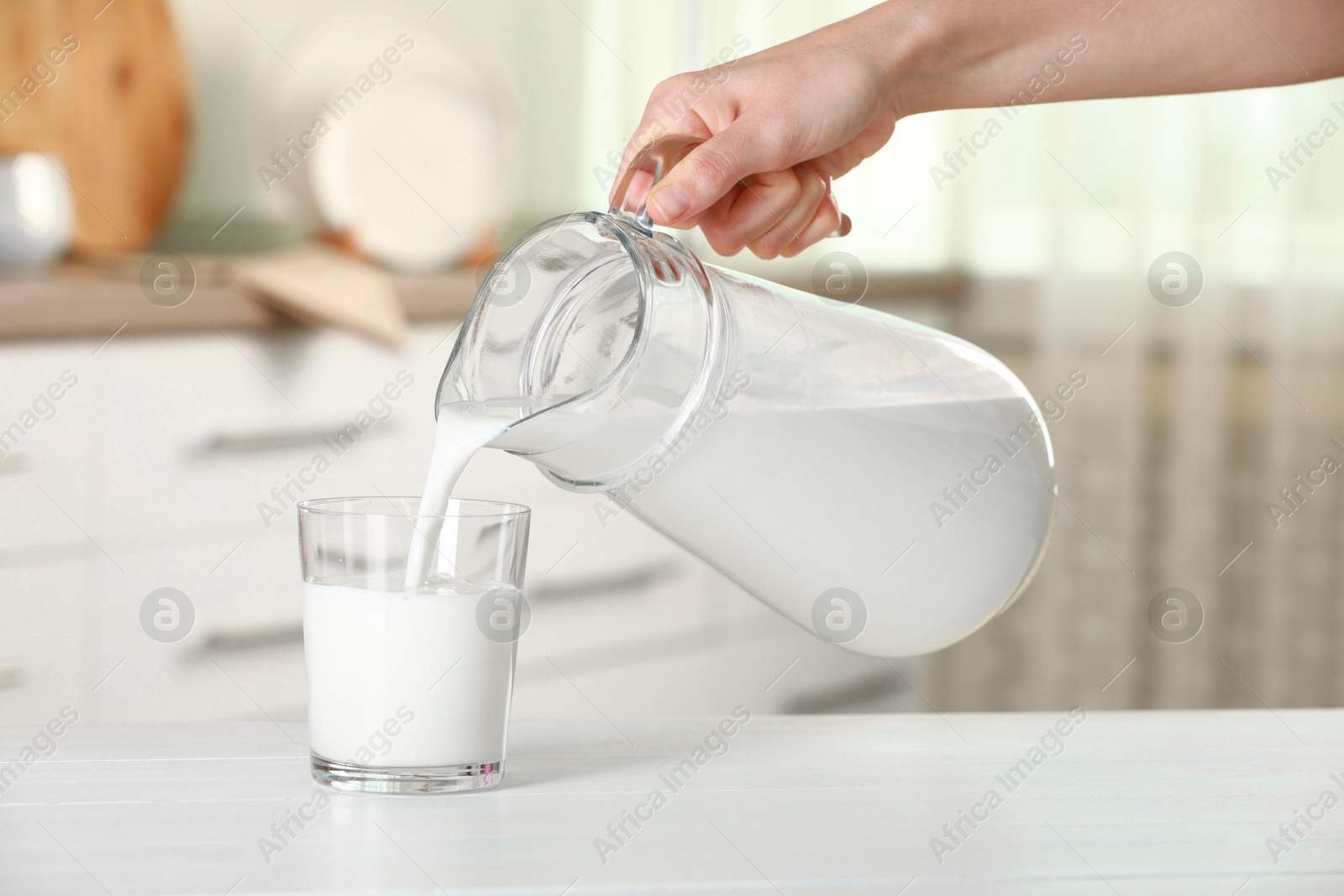 Photo of Woman pouring fresh milk from jug into glass at white wooden table indoors, closeup