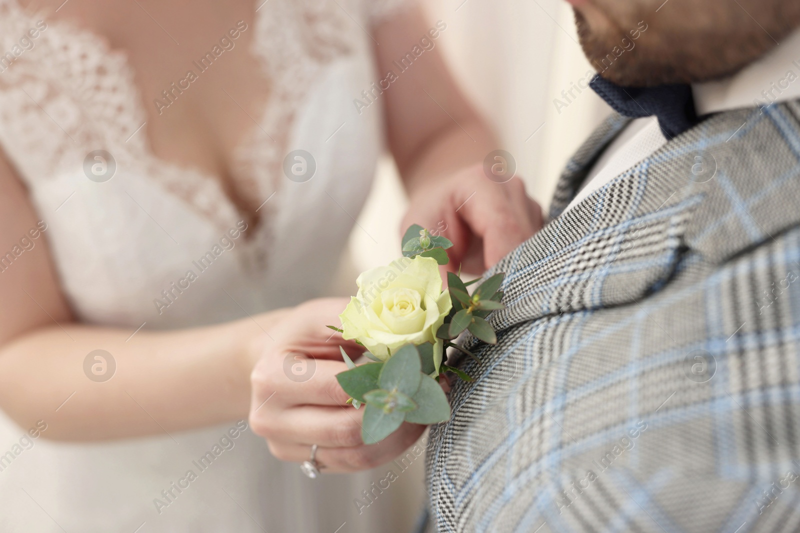 Photo of Bride putting boutonniere on her groom's jacket against light background, closeup. Wedding accessory