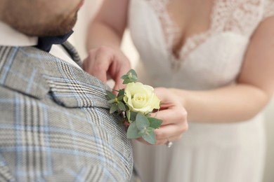Photo of Bride putting boutonniere on her groom's jacket against light background, closeup. Wedding accessory