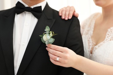 Bride putting boutonniere on her groom's jacket against light background, closeup. Wedding accessory