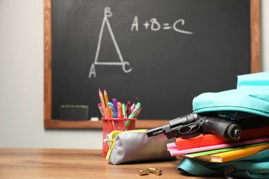 Gun, bullets and school stationery on wooden table near blackboard indoors, space for text