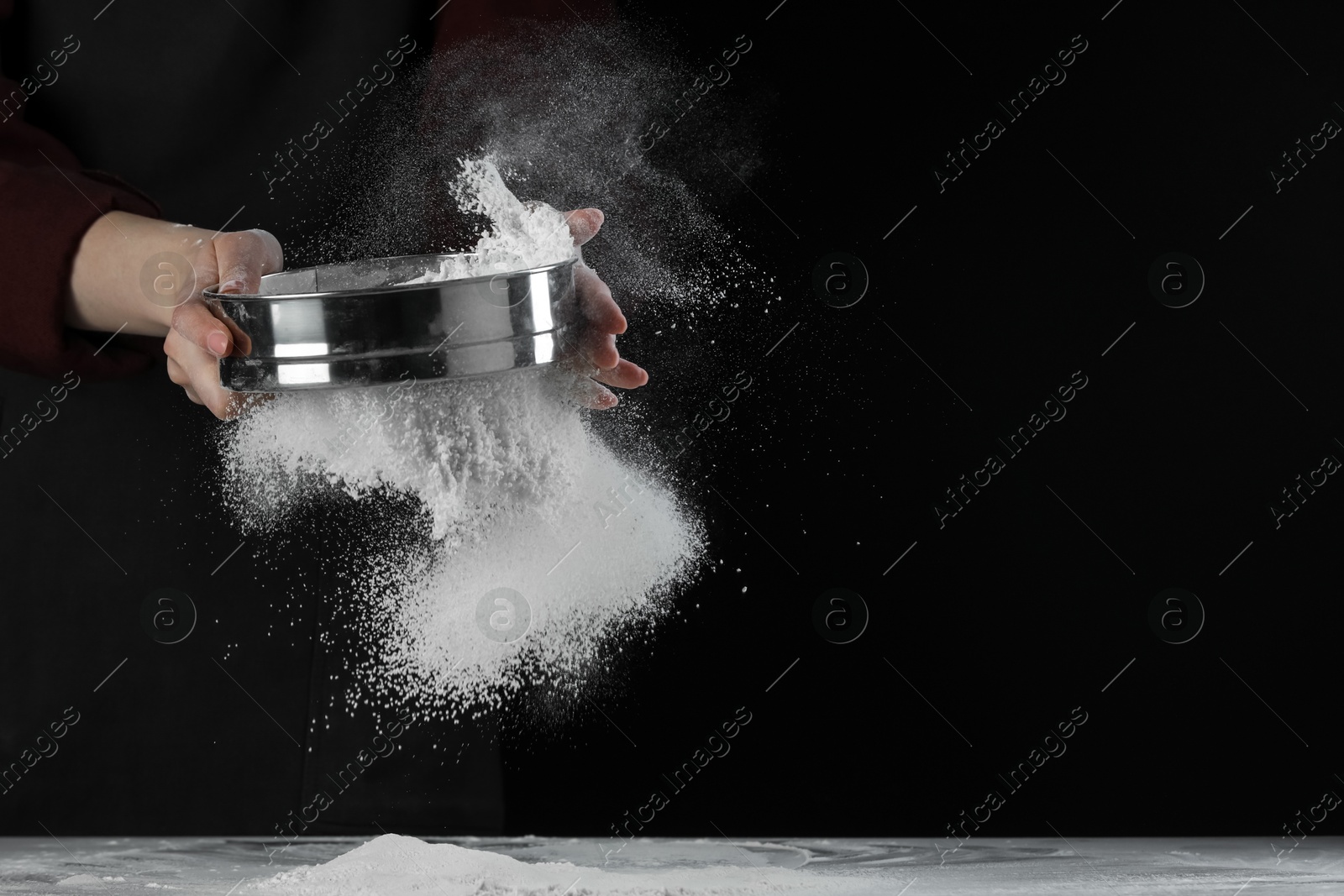 Photo of Woman sieving flour at table against black background, closeup