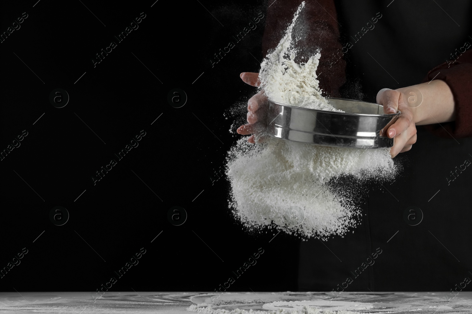 Photo of Woman sieving flour at table against black background, closeup