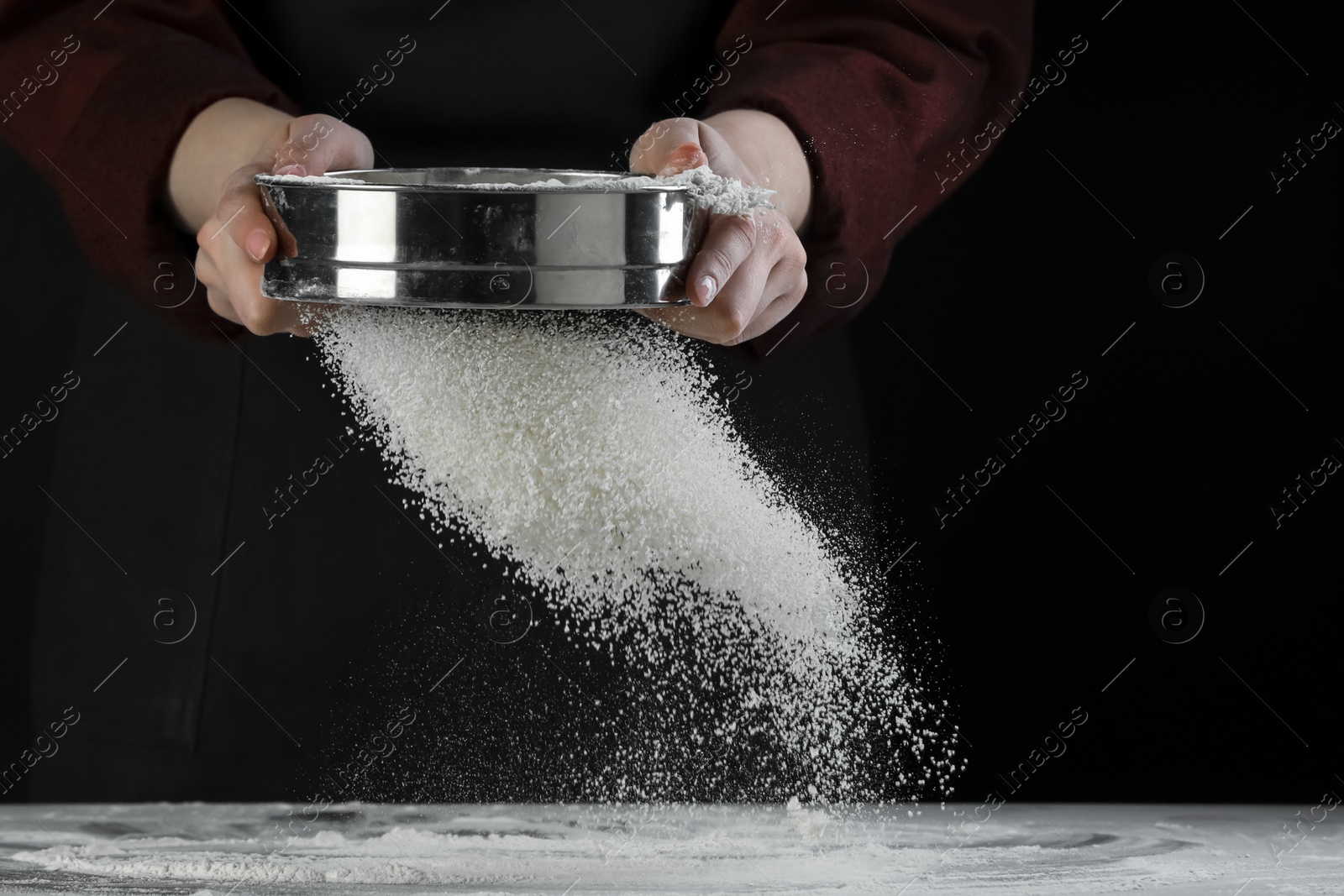 Photo of Woman sieving flour at table against black background, closeup