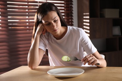 Photo of Eating disorder. Sad woman with fork and slice of cucumber at wooden table