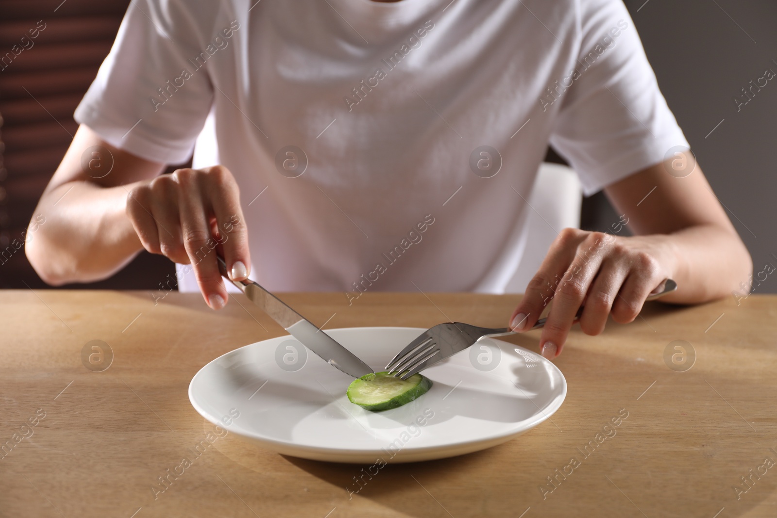 Photo of Eating disorder. Woman cutting cucumber at wooden table indoors, closeup