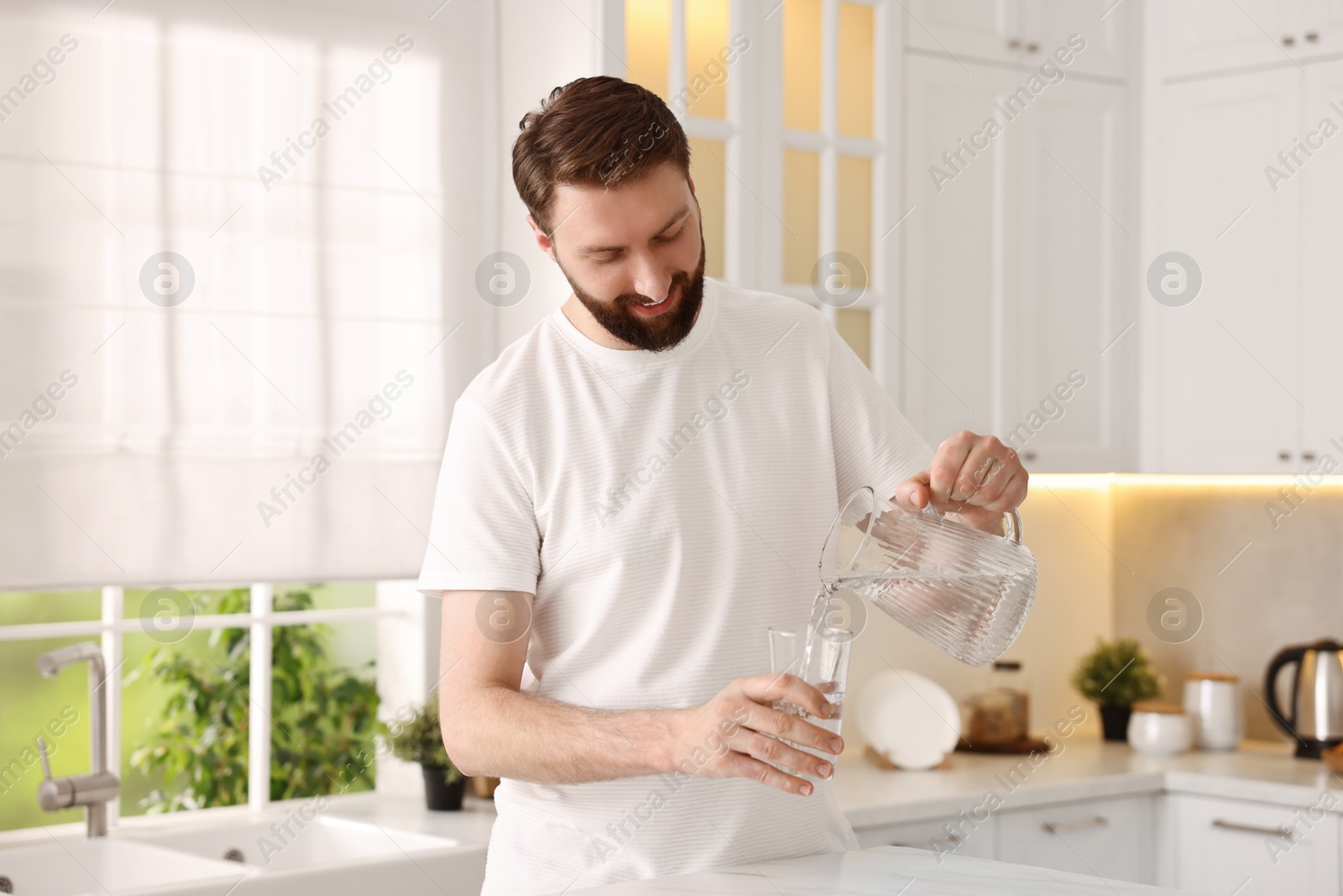 Photo of Happy young man pouring water into glass in kitchen at morning