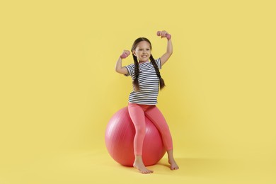 Photo of Cute little girl with dumbbells and fit ball on yellow background