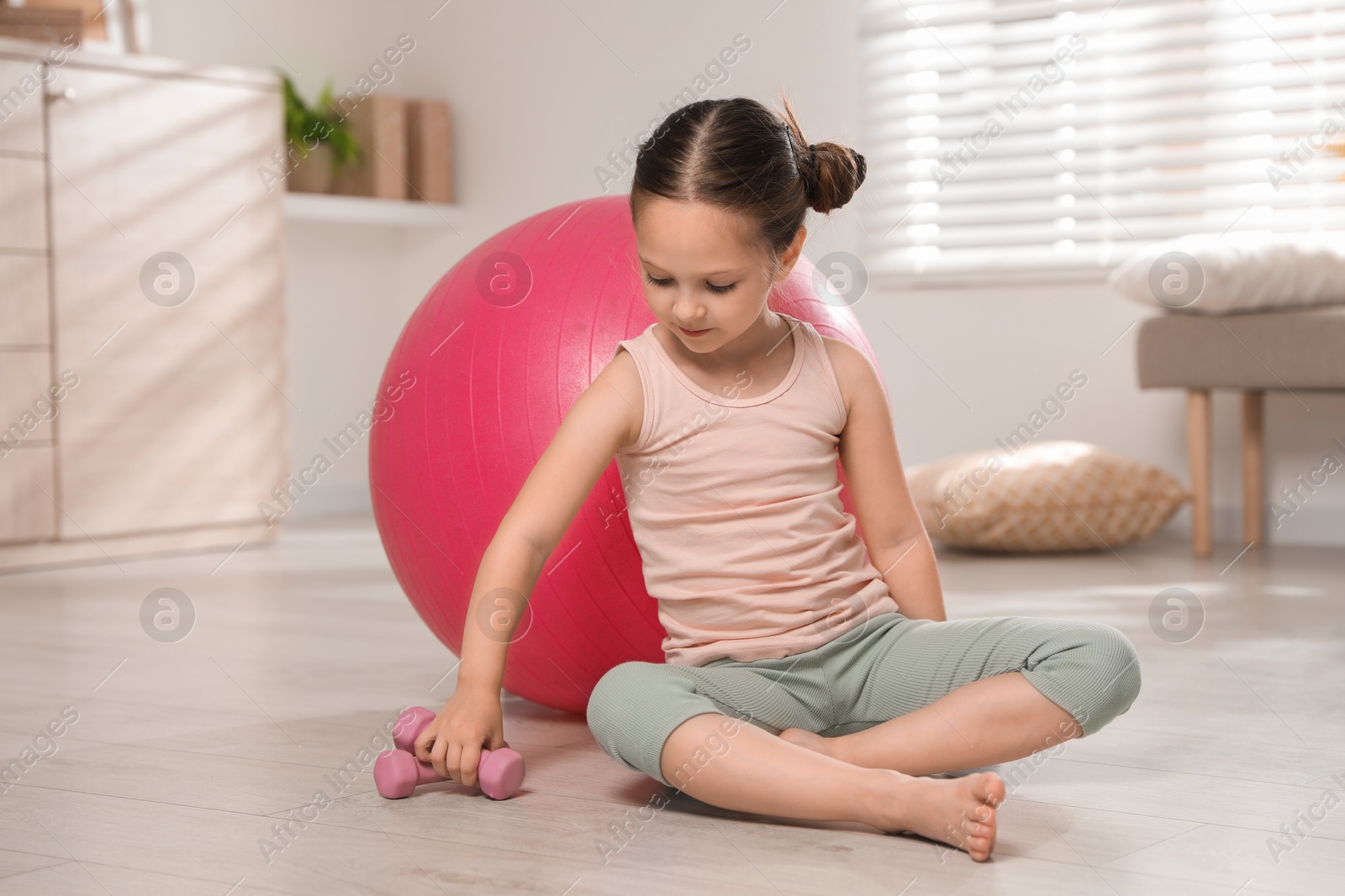 Photo of Cute little girl with dumbbells near fit ball at home