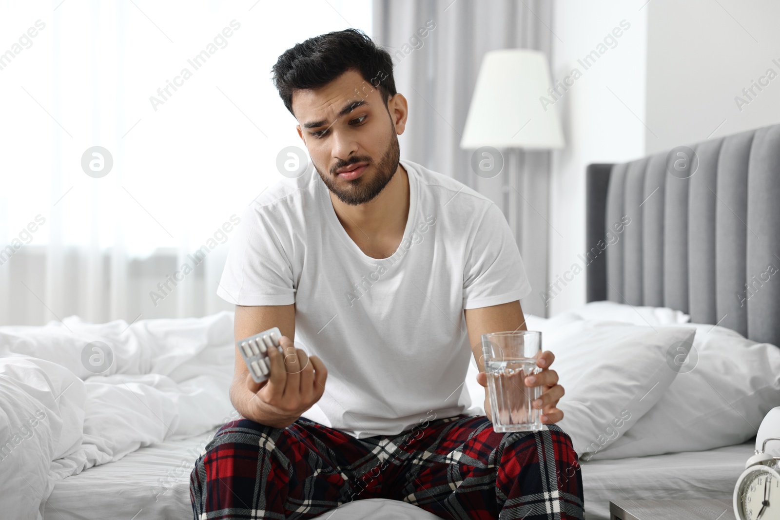 Photo of Man suffering from headache with glass of water and pills on bed at morning