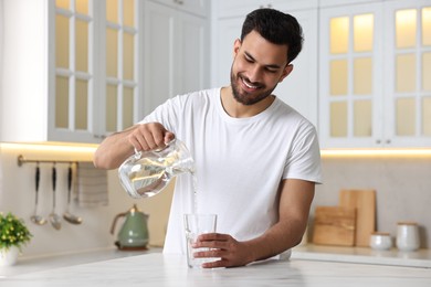 Photo of Morning of happy man pouring water from jug into glass at table in kitchen