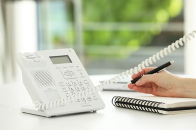 Assistant with telephone handset writing at white table against blurred green background, closeup