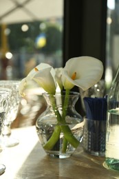 Photo of Beautiful calla lily flowers in glass vase on wooden table indoors, closeup