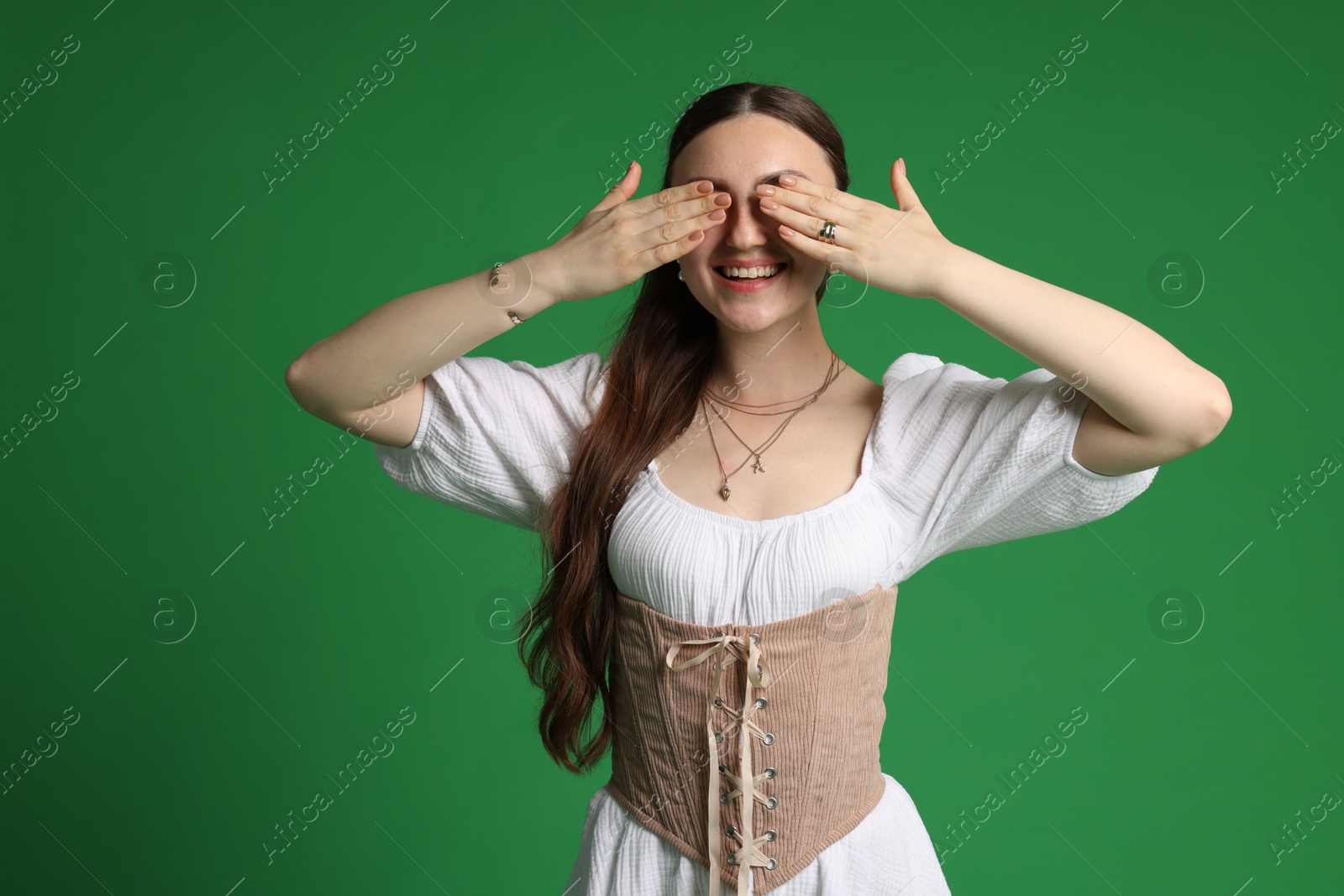 Photo of Smiling woman in stylish corset covering eyes with hands on green background