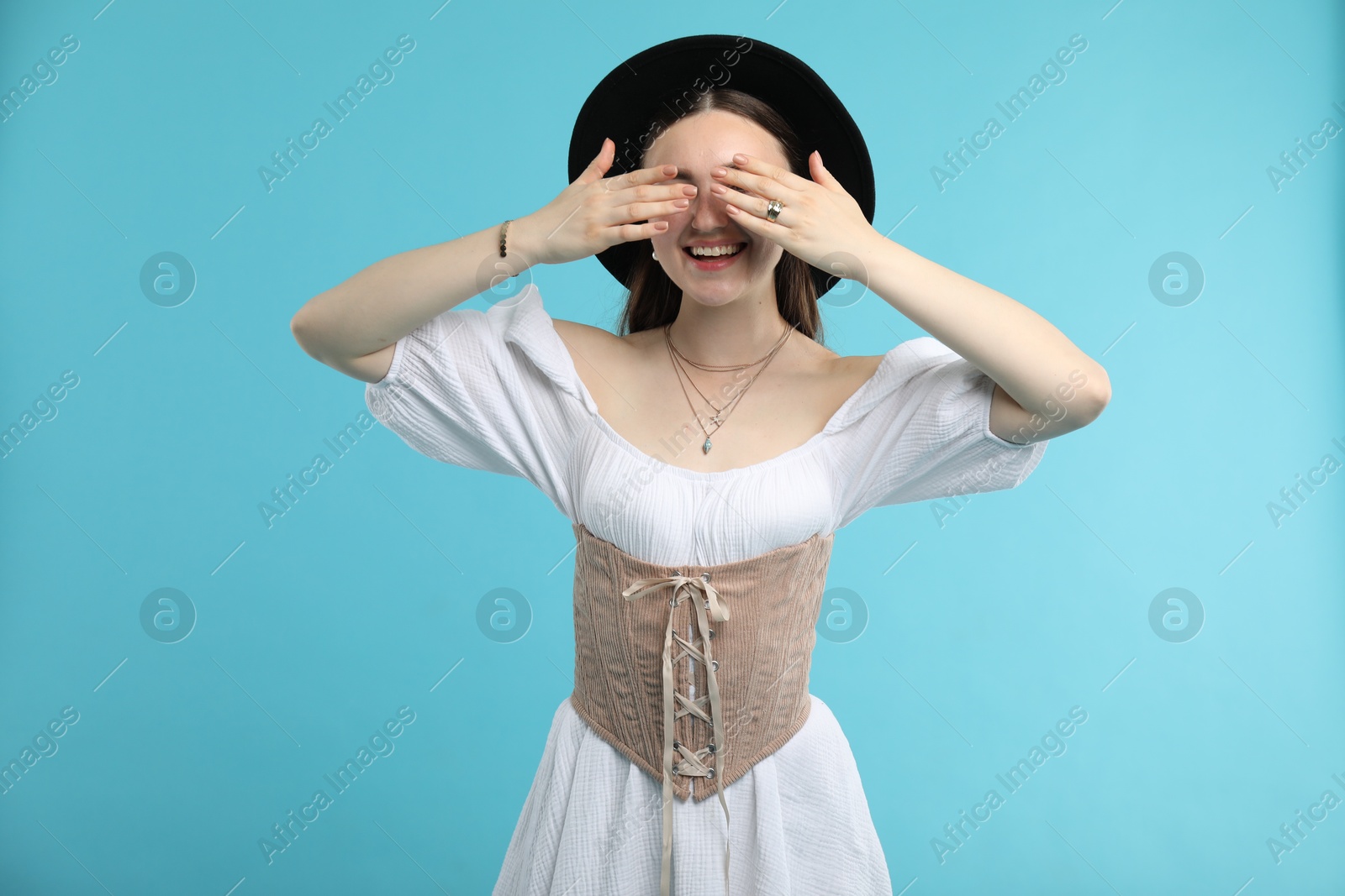 Photo of Smiling woman in velvet corset covering eyes with hands on light blue background