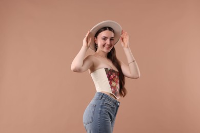 Smiling woman in stylish corset and hat posing on beige background