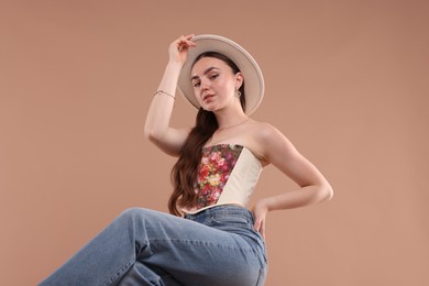 Photo of Beautiful woman in stylish corset and hat posing against beige background, low angle view