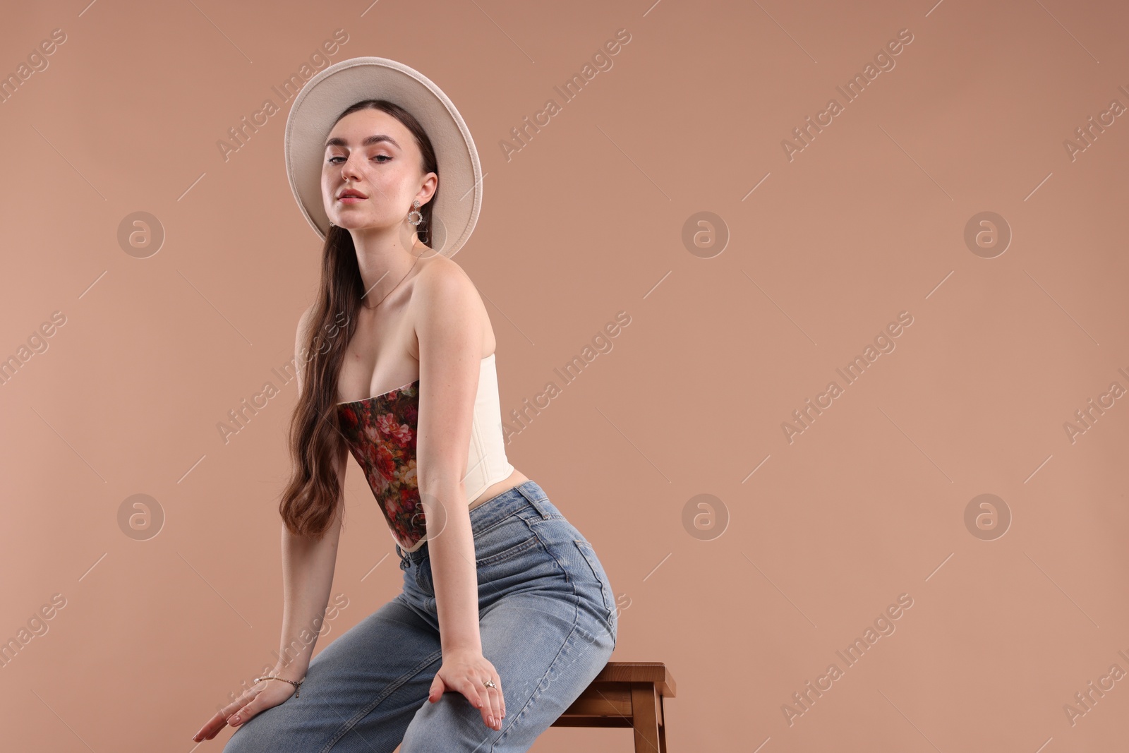 Photo of Beautiful woman in stylish corset and hat posing on stool against beige background. Space for text