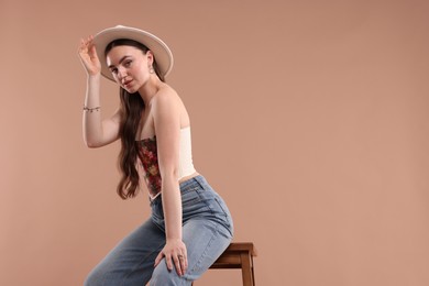 Beautiful woman in stylish corset and hat posing on stool against beige background. Space for text