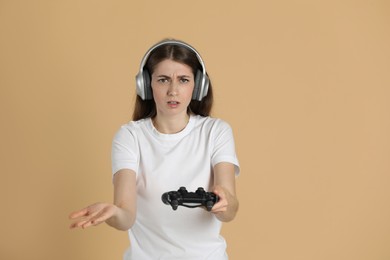 Unhappy woman in headphones with controller on beige background