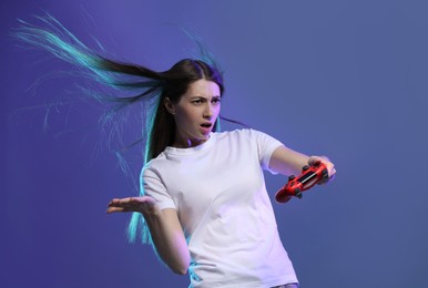 Surprised woman playing video games with controller on violet background, space for text