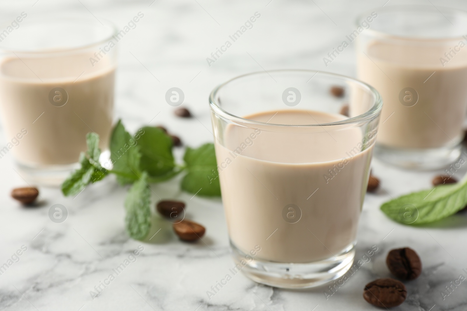Photo of Coffee cream liqueur in glasses, mint and beans on white marble table, closeup