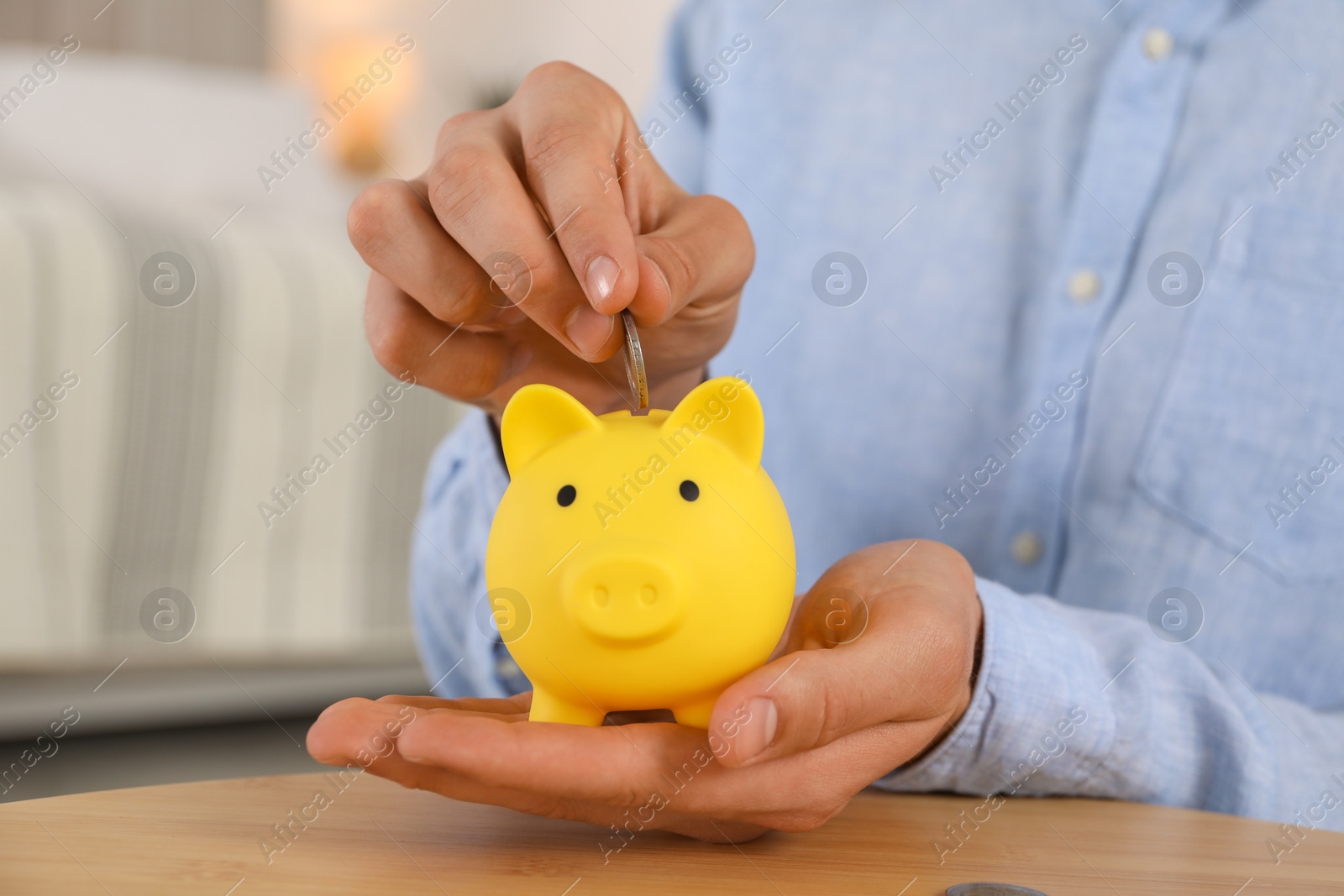 Photo of Man putting coin into yellow piggy bank on wooden table, closeup