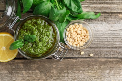 Tasty pesto sauce in jar, pine nuts, basil and lemon on wooden table, top view. Space for text