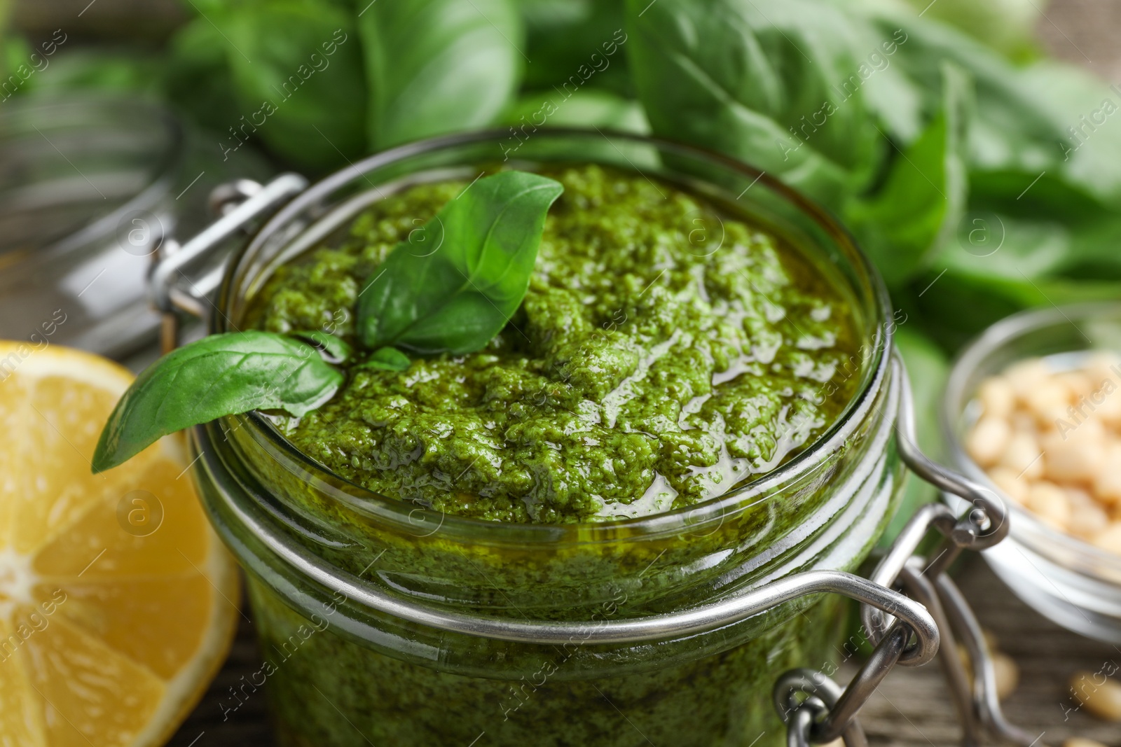 Photo of Tasty pesto sauce in glass jar, basil and lemon on table, closeup