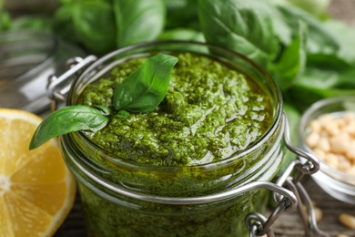 Tasty pesto sauce in glass jar, basil and lemon on table, closeup