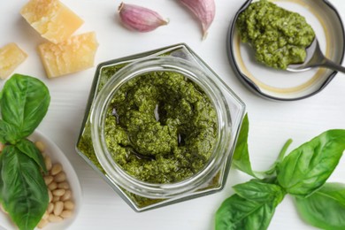 Photo of Tasty pesto sauce in jar, spoon, pine nuts, garlic and basil on white wooden table, flat lay