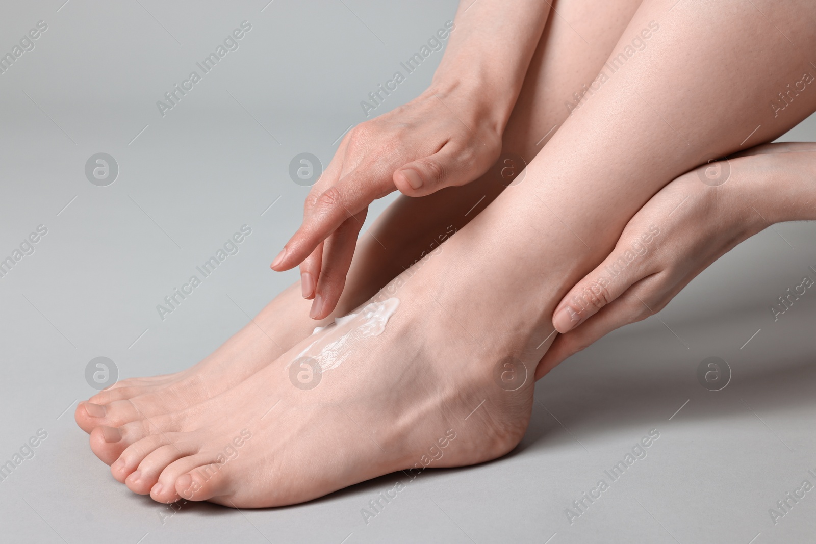 Photo of Woman applying moisturizing cream onto her feet on grey background, closeup. Body care