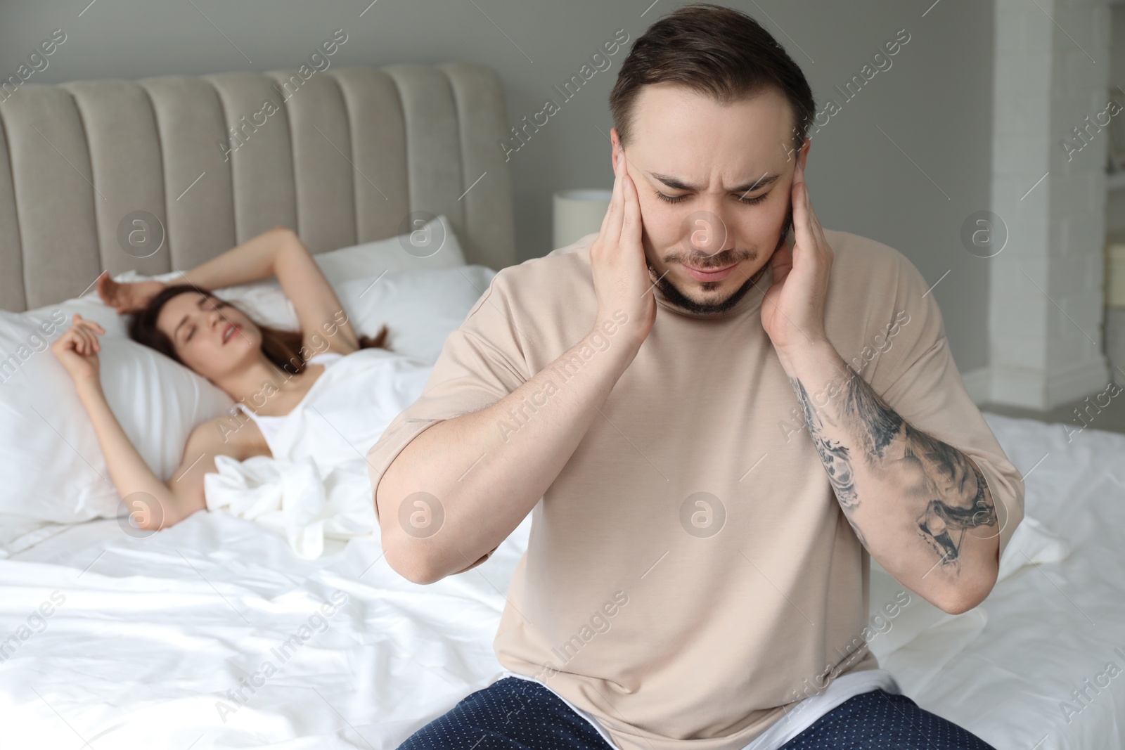 Photo of Bedtime. Irritated man covering ears near his snoring wife in bed at home