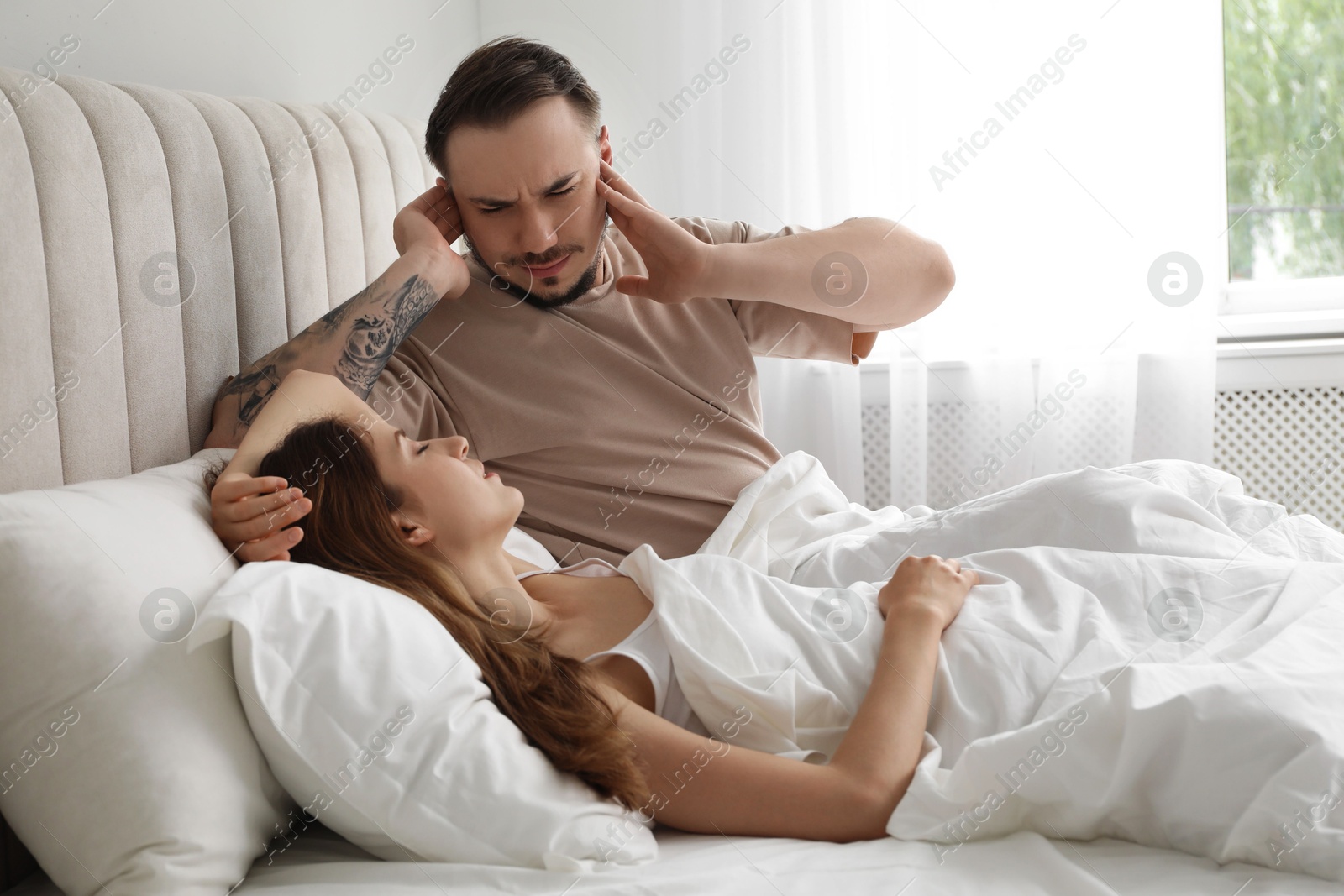 Photo of Bedtime. Irritated man covering ears near his snoring wife in bed at home