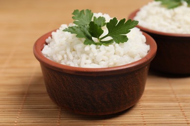 Photo of Bowls of delicious boiled rice with parsley on bamboo mat, closeup