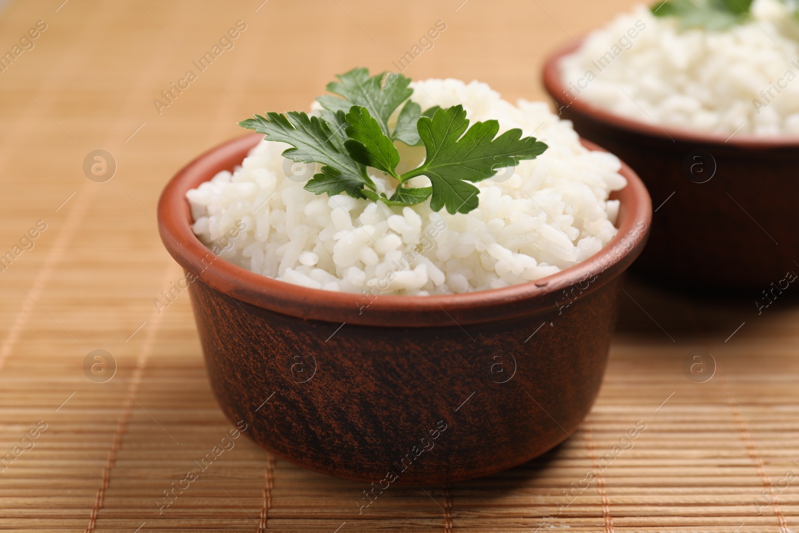 Photo of Bowls of delicious boiled rice with parsley on bamboo mat, closeup