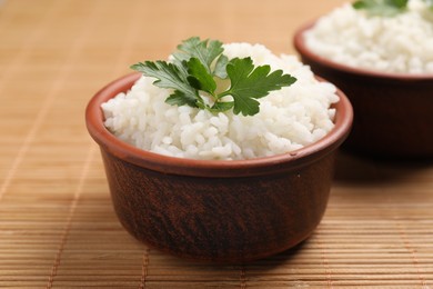 Photo of Bowls of delicious boiled rice with parsley on bamboo mat, closeup