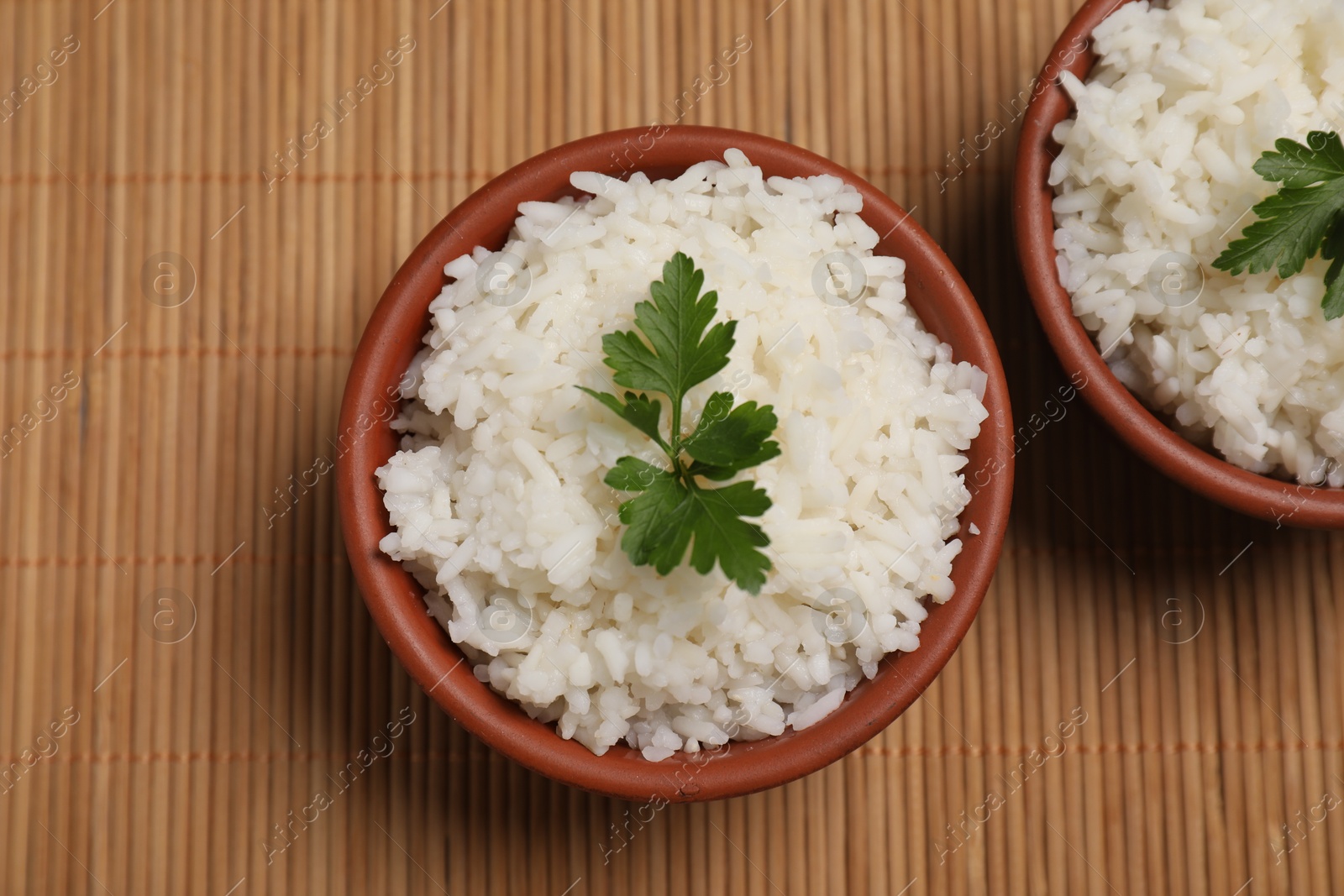 Photo of Bowls of delicious boiled rice with parsley on bamboo mat, top view