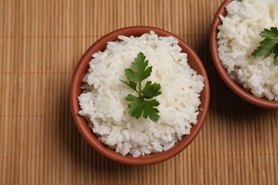 Photo of Bowls of delicious boiled rice with parsley on bamboo mat, top view